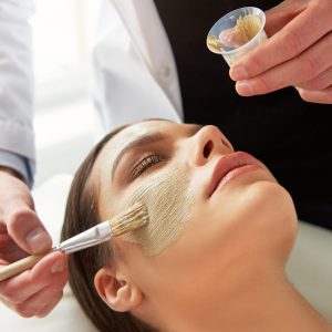Male cosmetologist putting mask on young female face with a brush during cosmetic procedure.